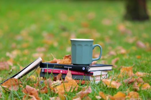 A serene autumn setup of books and a cup on grassy field with fallen leaves.